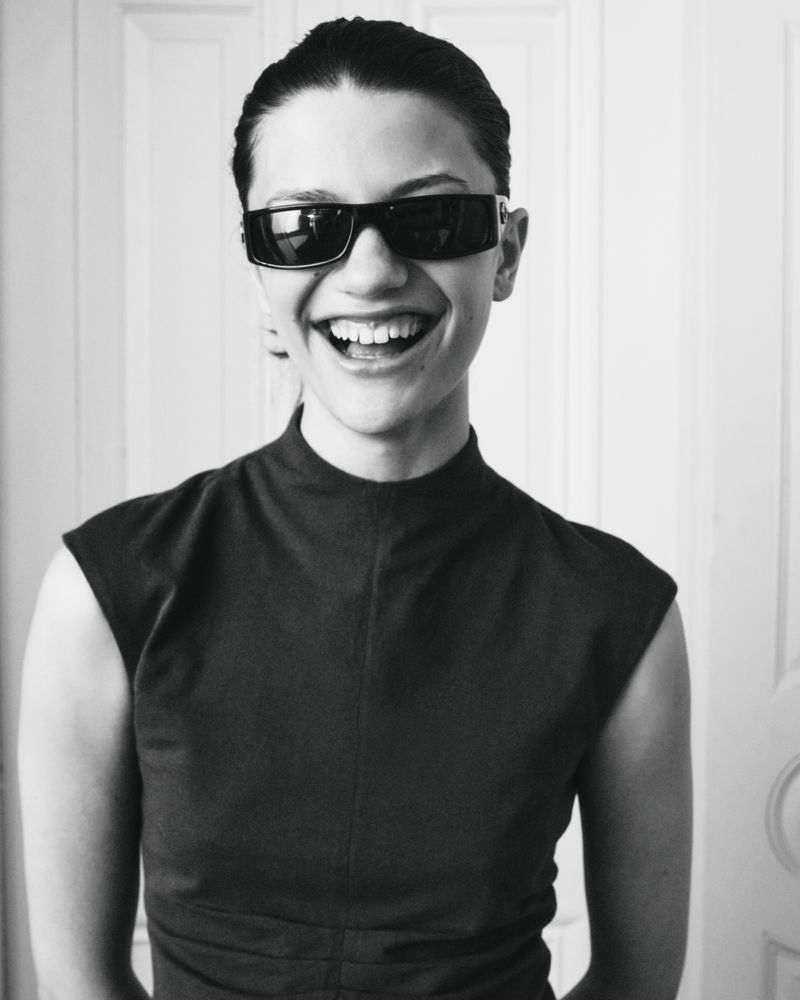 Smiling woman wearing dark rectangular sunglasses and a sleeveless high-neck black top in a black and white portrait.