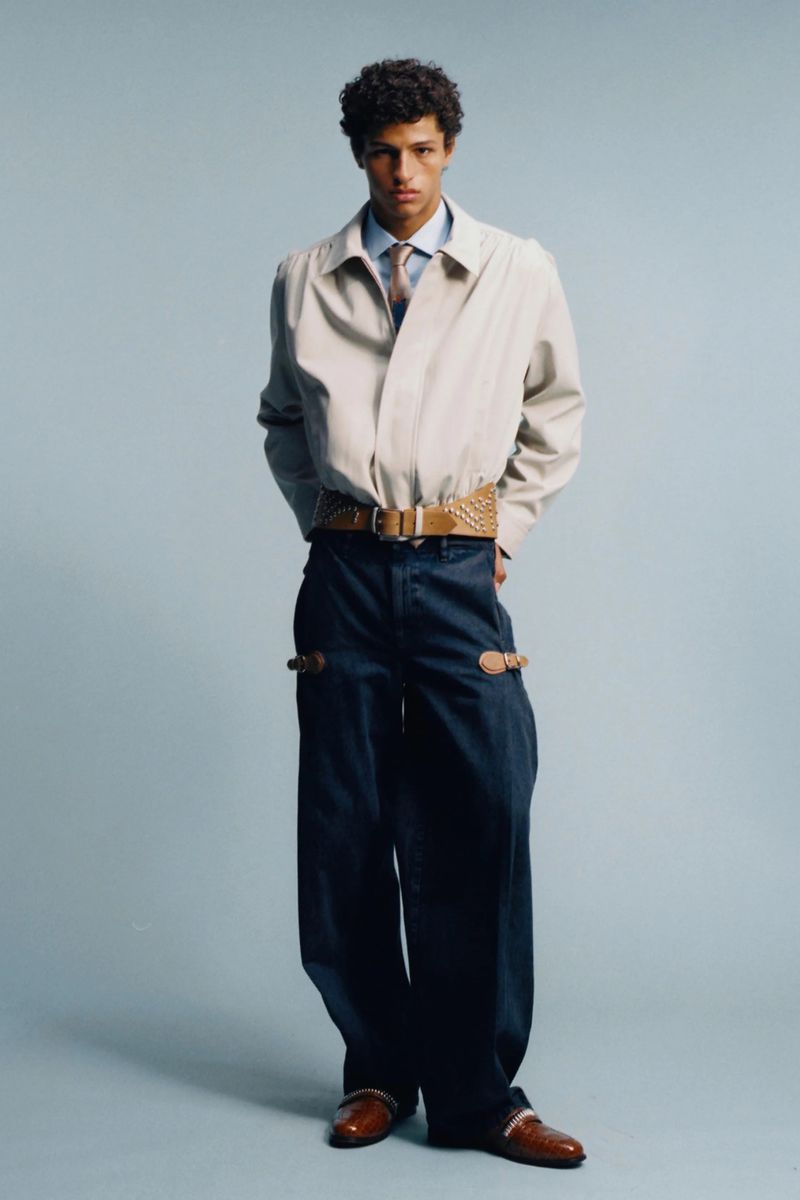 Model Adam Gerphagnon poses in a beige collared jacket, wide-leg trousers, and a wide belt against a light blue seamless background.