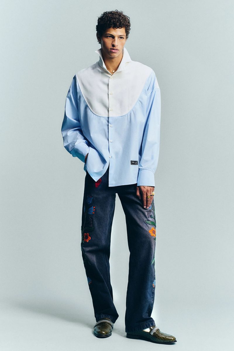 Model wearing a light blue two-tone shirt and dark floral embroidered jeans standing against a light grey studio background.