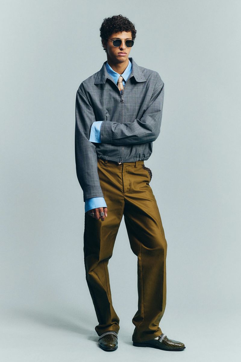 Model wearing a grey jacket over a blue shirt and brown trousers against a neutral background