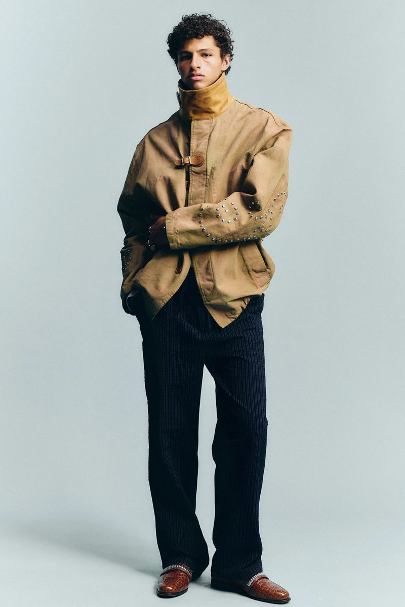 Model Adam Gerphagnon in a tan studded jacket and dark wide-leg trousers posing against a grey studio backdrop.