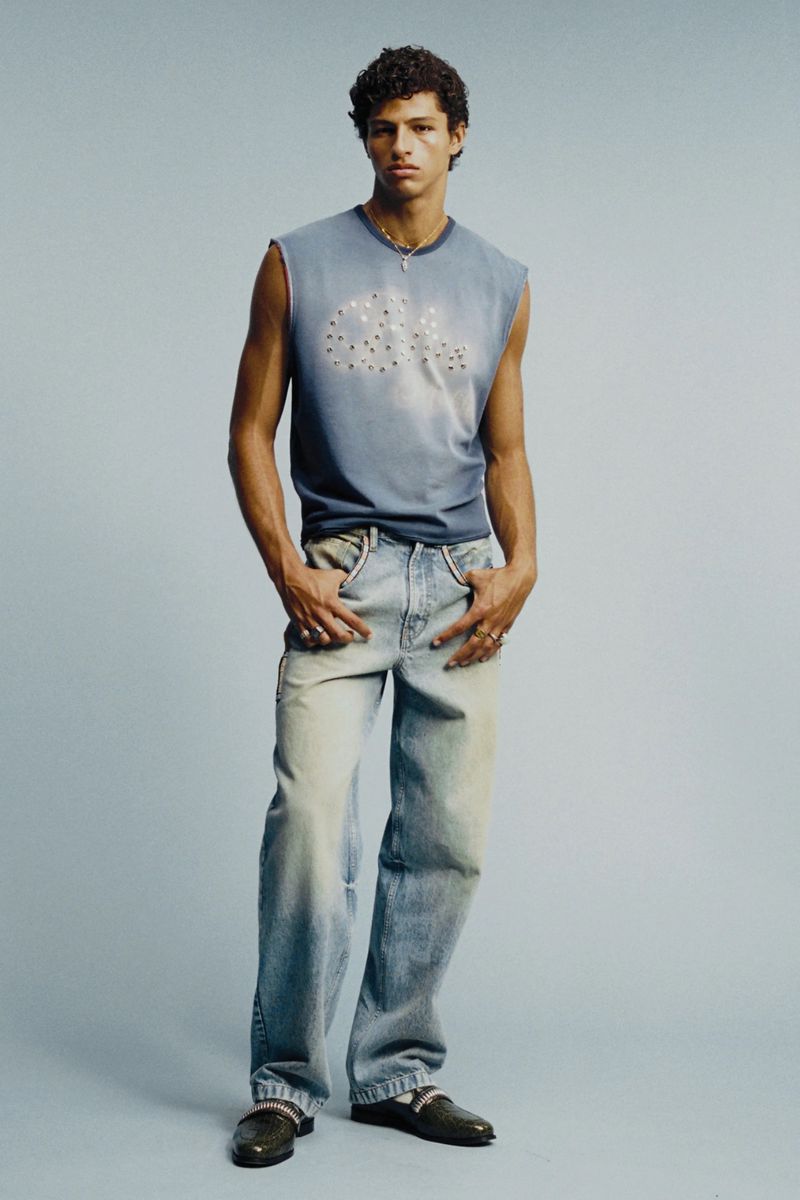 Model Adam Gerphagnon poses in a blue sleeveless graphic top and light wash denim jeans against a pale blue studio background.