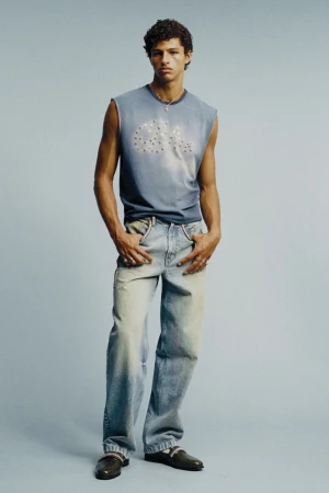 Model Adam Gerphagnon poses in a blue sleeveless graphic top and light wash denim jeans against a pale blue studio background.