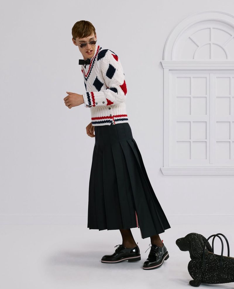 Fashionable adult wears patterned sweater and pleated skirt in studio setting, paired with black oxfords and dachshund bag.