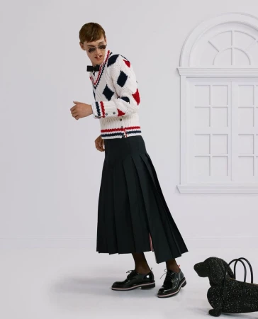 Fashionable adult wears patterned sweater and pleated skirt in studio setting, paired with black oxfords and dachshund bag.