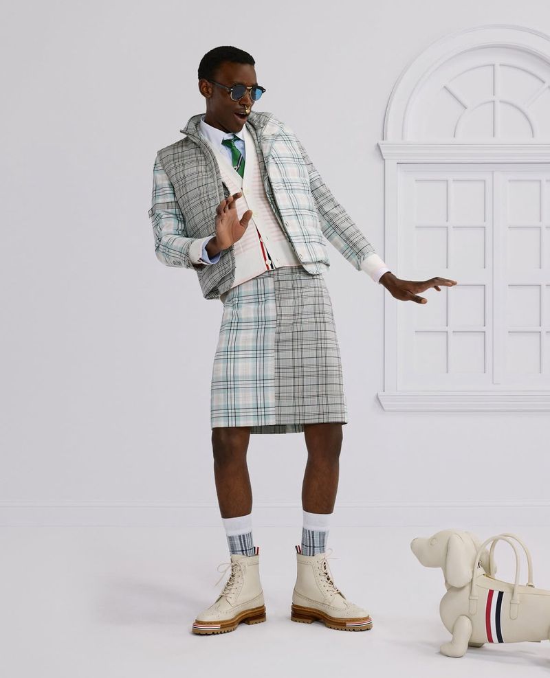 Man in plaid suit jacket and kilt with dog-shaped bag in a bright studio. He wears sunglasses and boots, striking a pose.