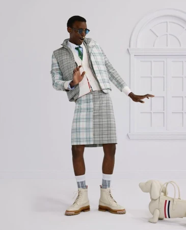 Man in plaid suit jacket and kilt with dog-shaped bag in a bright studio. He wears sunglasses and boots, striking a pose.