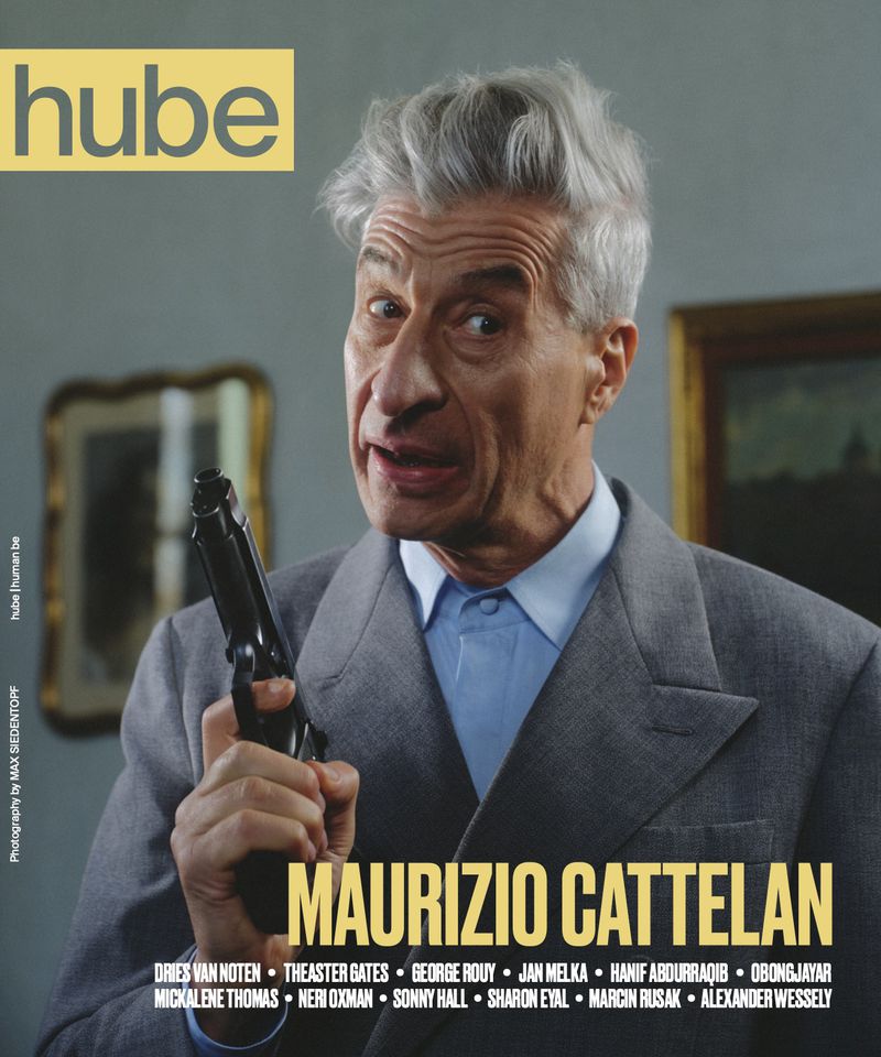 Artist Maurizio Cattelan holding a handgun wearing a grey suit jacket and light blue shirt on a magazine cover.