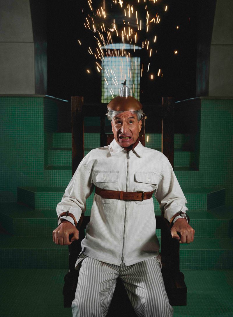 Maurizio Cattelan strapped into an electric chair with sparks flying from his helmet for Hube Magazine.