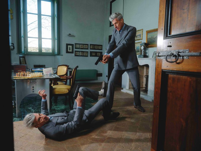 Artist Maurizio Cattelan confronts a duplicate of himself, both holding handguns in a room while dressed in grey suits.