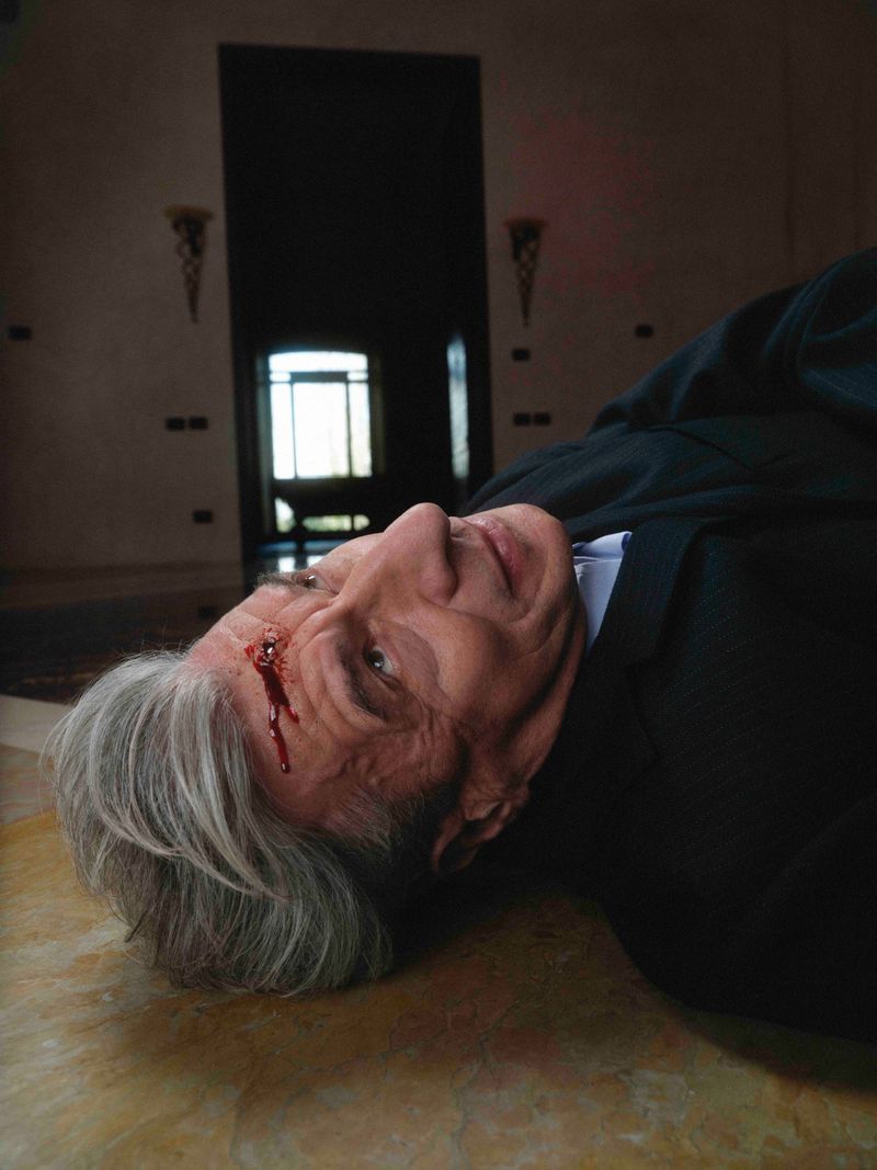 Artist Maurizio Cattelan lying on a floor wearing a dark suit with stage blood on his forehead for Hube Magazine.