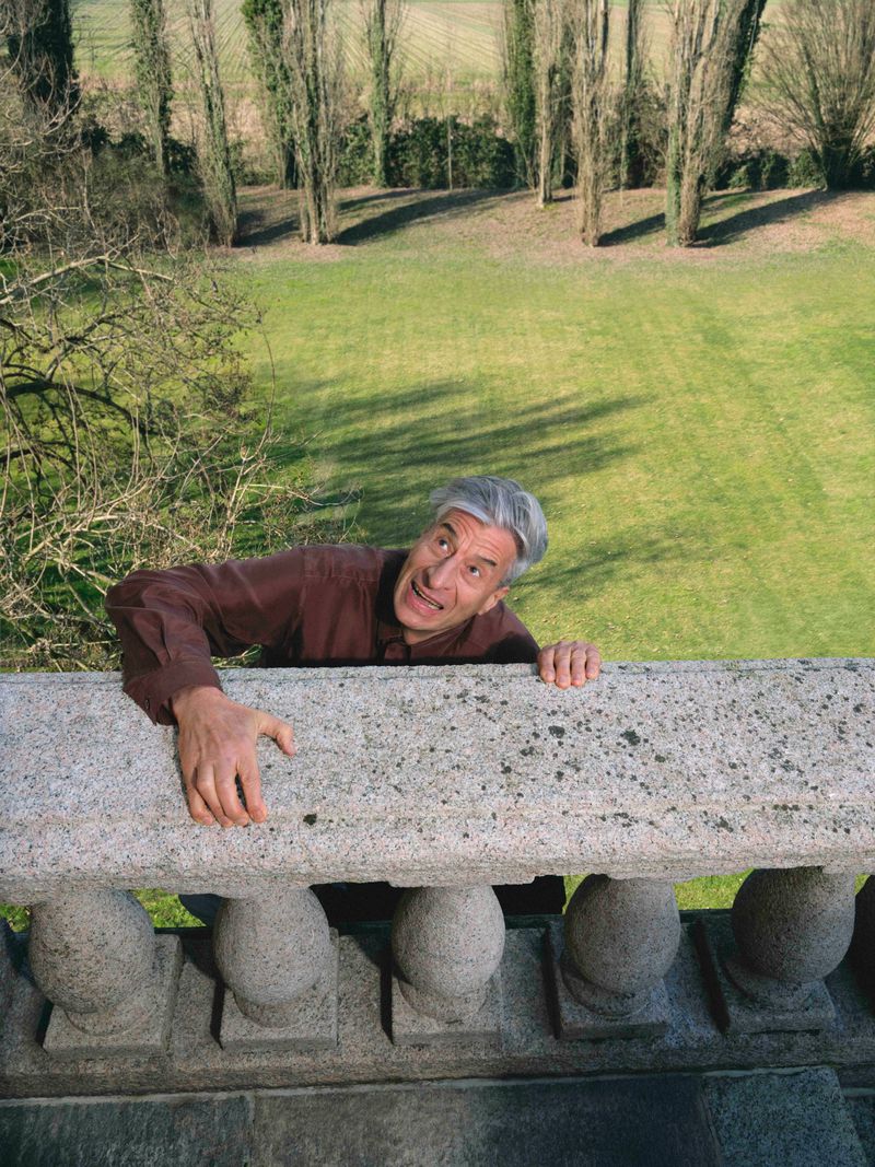 Artist Maurizio Cattelan hangs from a classical stone balustrade, looking up with an alarmed expression against a green lawn.