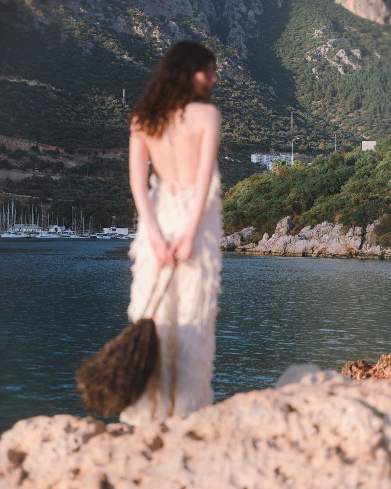 Model seen from behind in an off-white backless dress holding a brown fringe bag while standing on a rocky coastal cliff.