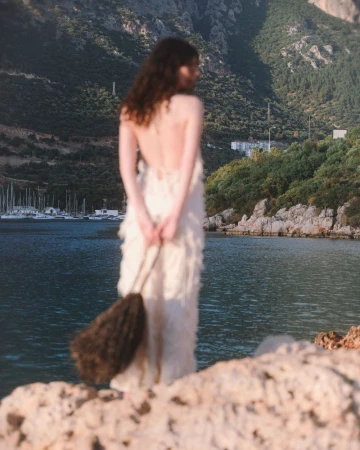Model seen from behind in an off-white backless dress holding a brown fringe bag while standing on a rocky coastal cliff.