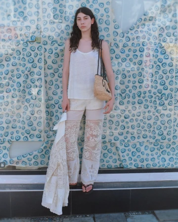 Model Oyku Bastas posing in white lace trousers, a tank top, and a woven raffia bag in front of an evil eye patterned backdrop.