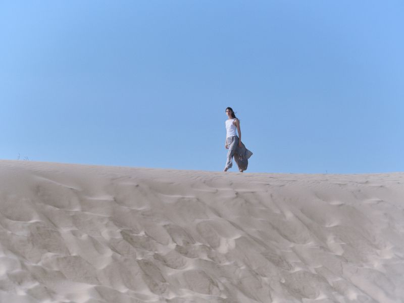 Model walking across a sandy dune under a clear blue sky for the BIMBA Y LOLA Cala Bimba campaign, photographed by Furkan Temir