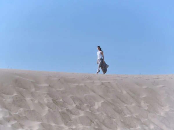 Model walking across a sandy dune under a clear blue sky for the BIMBA Y LOLA Cala Bimba campaign, photographed by Furkan Temir