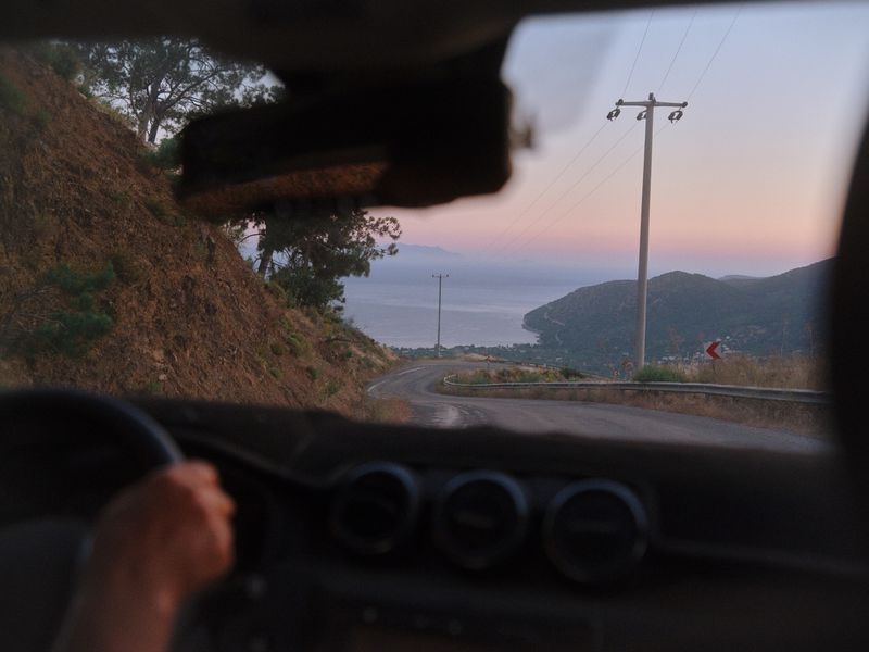 Point of view shot from inside a car driving along a winding coastal mountain road toward the sea at dusk.