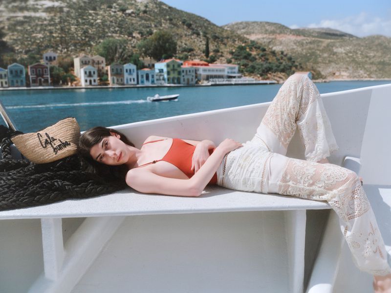 Model Oyku Bastas reclining on a white boat in an orange swimsuit and white lace trousers with a straw tote bag before a coastal bay.