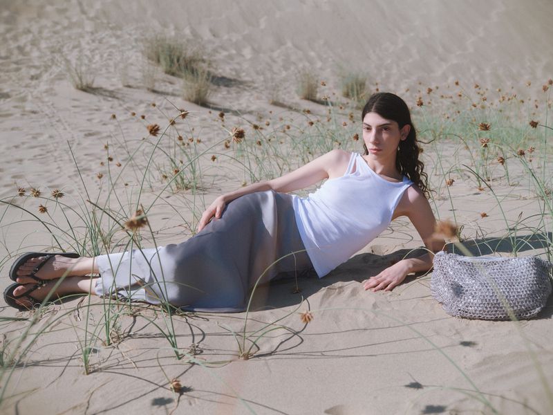 Model Oyku Bastas reclining on sand dunes in a white tank top and grey skirt beside a silver mesh bag for BIMBA Y LOLA.