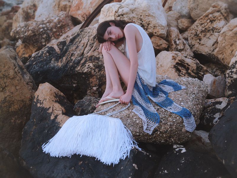 Model Oyku Bastas rests on large coastal rocks wearing a white sleeveless top with a large white fringe tote bag in the foreground.