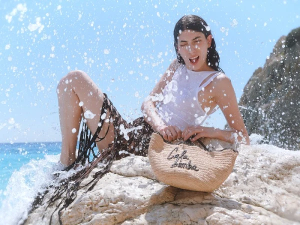 Model Oyku Bastas poses on coastal rocks amidst splashing water, holding a straw bag for BIMBA Y LOLA.