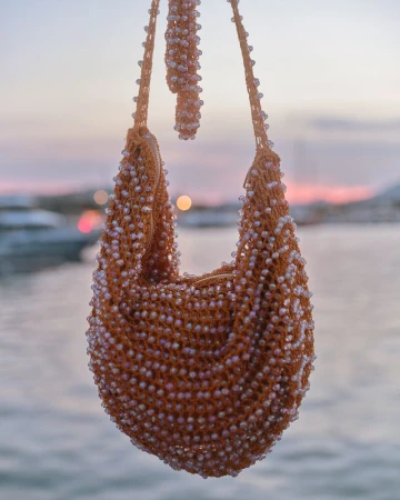 An orange crochet shoulder bag embellished with iridescent beads hanging against a soft-focus harbor sunset.