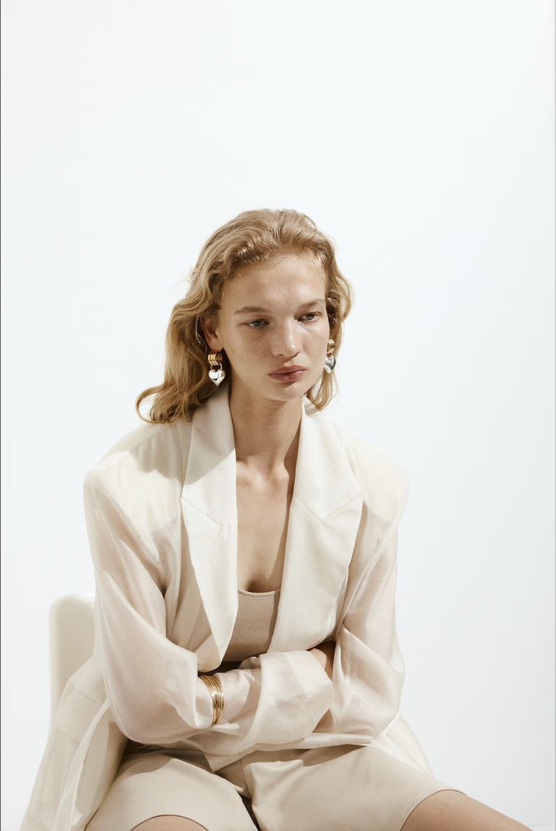Young woman in a beige suit sitting against a white background, looking down, with earrings and a bracelet. Indoor fashion shot.