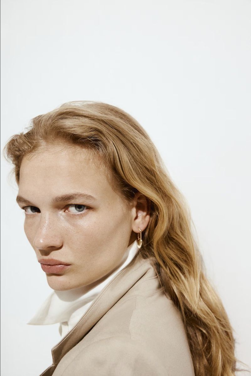 Blonde woman with blue eyes poses in a studio, wearing a beige coat and a white top, showcasing a gold earring against a white backdrop.