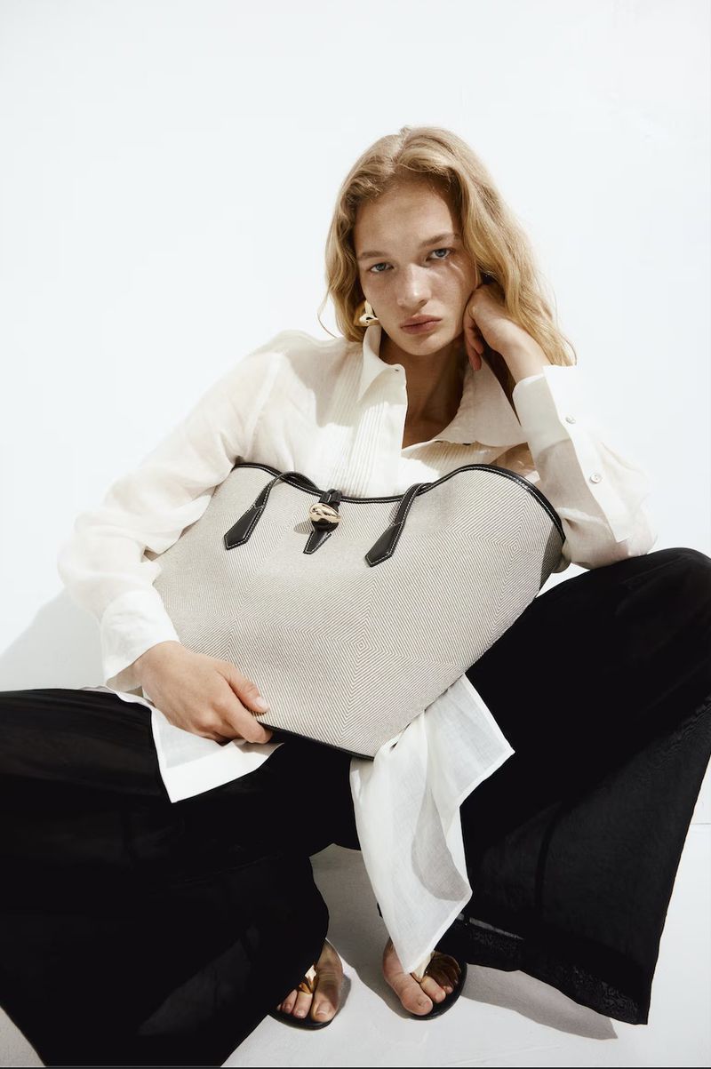 Studio shot of a fair-skinned adult female model with an open white button-down shirt holding a light-colored tote bag.