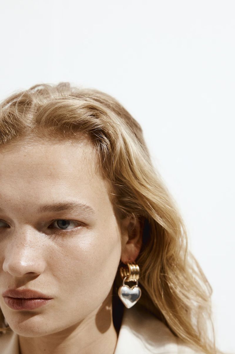 Close-up of a fair-skinned woman with blonde wavy hair, wearing a heart-shaped silver and gold earring against a bright white background.