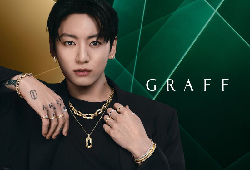 Jung Kook wearing gold chains and diamond rings from the Laurence Graff Signature collection against an emerald green background.