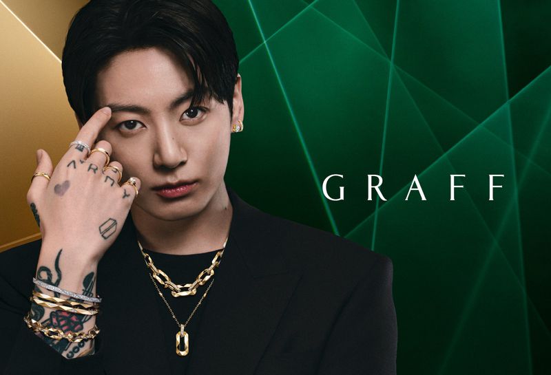 Jung Kook wearing layered gold Graff jewelry, a black blazer, and displaying hand tattoos against a dark green graphic backdrop.