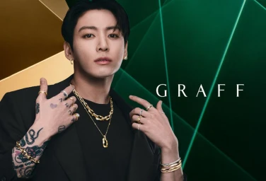Jung Kook wearing layered gold necklaces, rings, and bracelets in a black blazer against a deep green Graff backdrop.