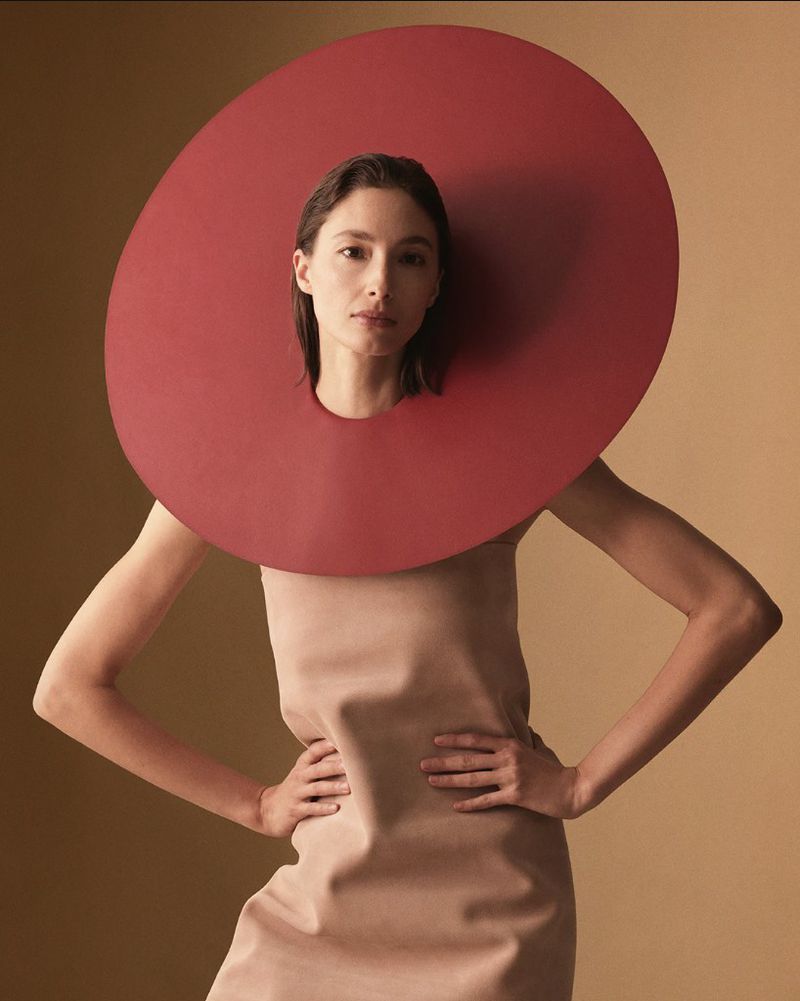 Alexandra Agoston poses with hands on hips wearing a nude dress and an oversized circular red collar against a tan backdrop.