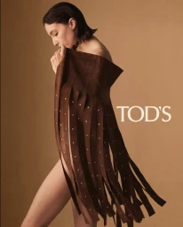 Model Alexandra Agoston poses in profile holding a brown studded suede fringe cape against a warm studio backdrop for Tod's.
