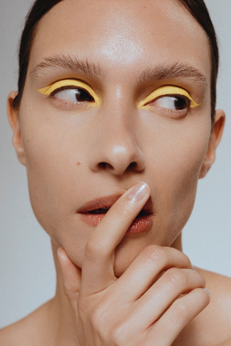 Close up portrait of a model with vibrant yellow eyeliner and finger touching lips against a plain background