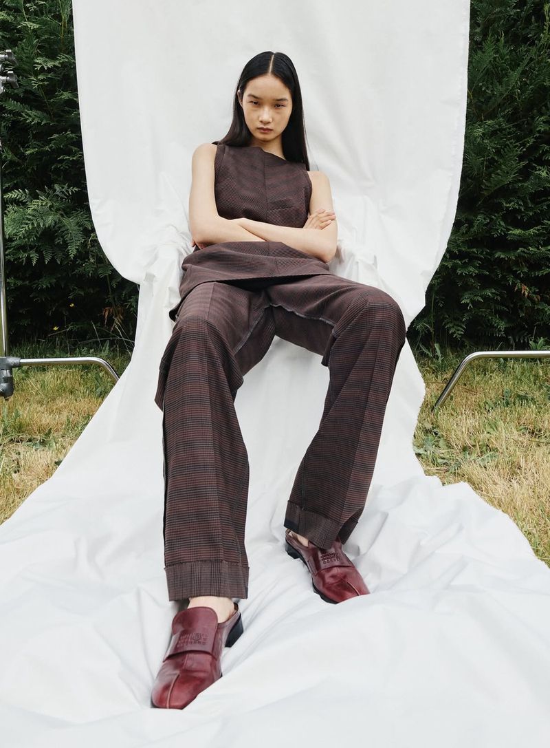 Asian woman in fashionable brown outfit poses in a seated position on a white fabric, set against a background of green foliage and grass.
