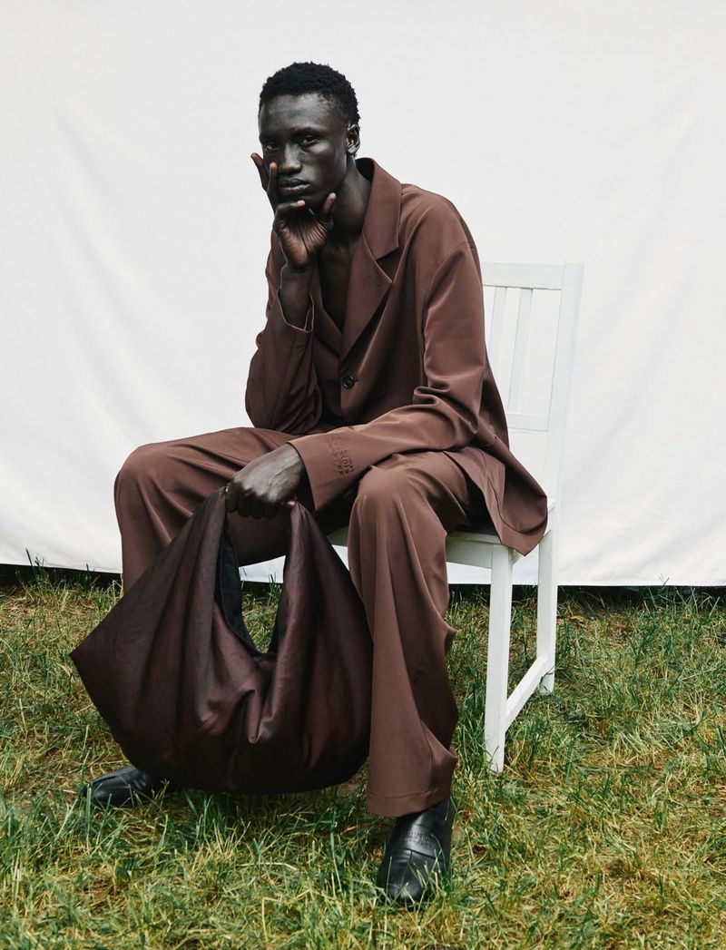 Fashion shot of a dark-skinned male model wearing a brown suit, holding a large unstructured bag, sitting on a white chair on grass.