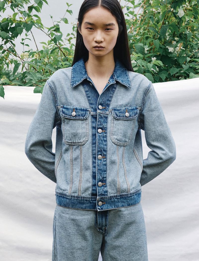 A young Asian woman is posing in a medium shot, wearing a denim jacket and jeans against a backdrop of greenery and a neutral background.
