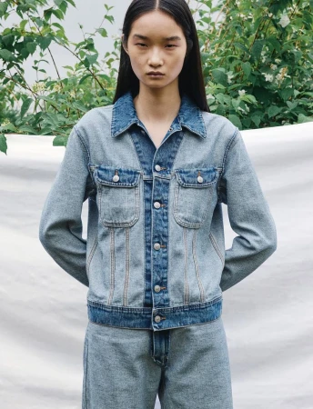 A young Asian woman is posing in a medium shot, wearing a denim jacket and jeans against a backdrop of greenery and a neutral background.