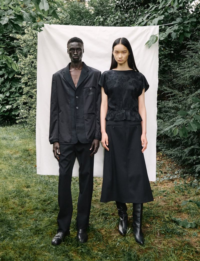 A duo of models, an adult man and woman, pose in a verdant outdoor setting against a white backdrop, both attired in black fashion pieces.