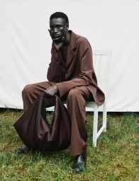 Black man wearing a brown suit holding a large dark handbag while seated on a white chair outdoors.