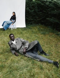 Black man reclining on grass wearing a grey trench coat with a model seated on a white chair before a backdrop in a forest