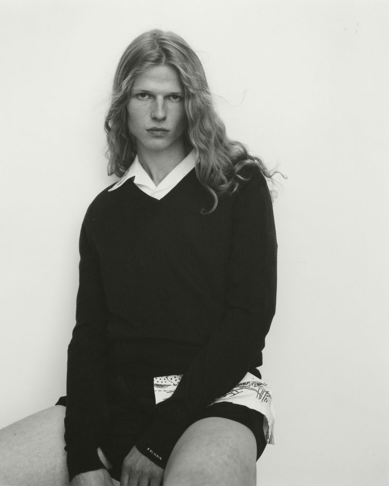 A model with long wavy hair wearing a black v-neck sweater over a white collared shirt, seated against a plain pale background.