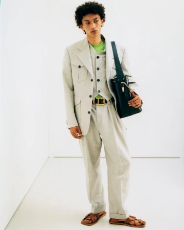 A male model in a pale grey three-piece tailored safari suit, lime green shirt, and brown sandals standing in a white studio.