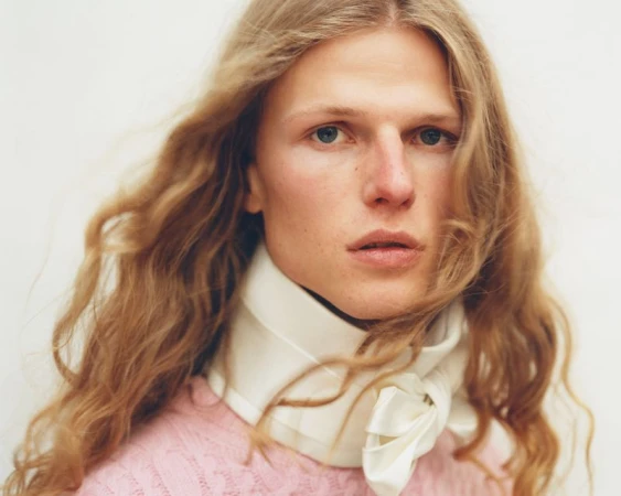 Close-up portrait of a model with long wavy blond hair wearing a white structured neck scarf and a light pink knit sweater.