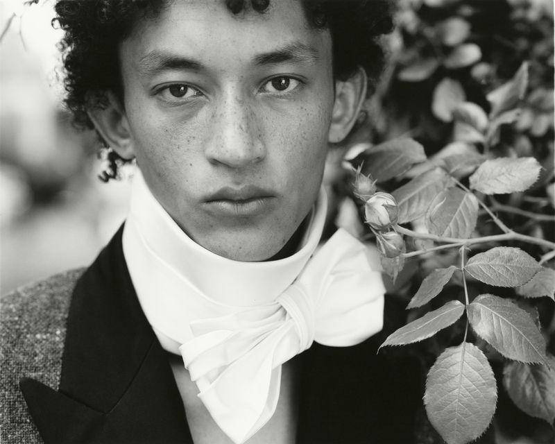 Monochrome close-up portrait of a young man wearing an oversized white neck bow collar beside leafy rose branches.