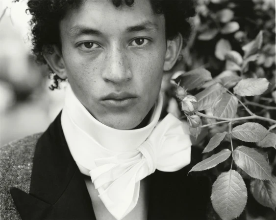 Monochrome close-up portrait of a young man wearing an oversized white neck bow collar beside leafy rose branches.