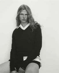 A model with long wavy hair wearing a black v-neck sweater over a white collared shirt, seated against a plain pale background.
